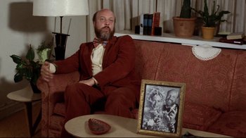 Movie still from “Eating Raoul” (1982), directed by Paul Bartel – A man sitting on a couch in front of a picture; Medium shot, Over the shoulder angle