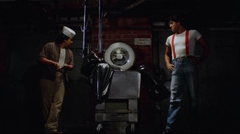 Movie still from “Eating Raoul” (1982), directed by Paul Bartel – Two men standing next to each other in front of a machine; Wide shot, Over the shoulder angle