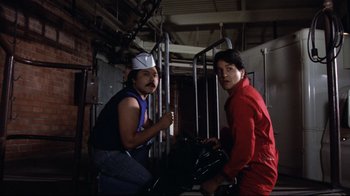 Movie still from “Eating Raoul” (1982), directed by Paul Bartel – Two men are posing for a picture in an industrial setting; Medium shot, Low angle