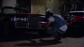 Movie still from “Eating Raoul” (1982), directed by Paul Bartel – A man squatting next to a car on the street; Wide shot, High angle