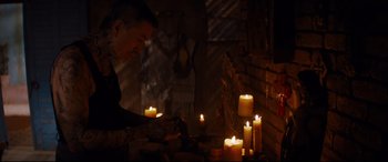 Movie still from “El Chicano” (2018), directed by Ben Hernandez Bray – A man is lighting candles in a dark room; Medium shot, Over the shoulder angle