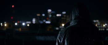 Movie still from “El Chicano” (2018), directed by Ben Hernandez Bray – A person wearing a hooded jacket standing in front of a city skyline at night; Close Up shot, Over the shoulder angle