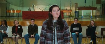 Movie still from “Election” (1999), directed by Alexander Payne – A woman standing in front of a microphone in front of a group of people; Medium shot, Over the shoulder angle