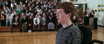 Movie still from “Election” (1999), directed by Alexander Payne – A man standing in front of a crowd of people in a gym; Medium shot, Over the shoulder angle