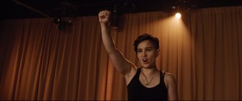 Movie still from “Dumplin'” (2018), directed by Anne Fletcher – A person raising their fist in the air in front of a curtain; Medium shot, Low angle