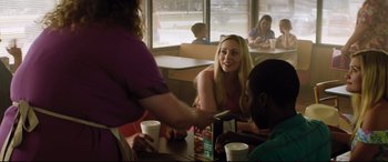Movie still from “Dumplin'” (2018), directed by Anne Fletcher – A woman is smiling at a table with two other people; Medium shot, Over the shoulder angle