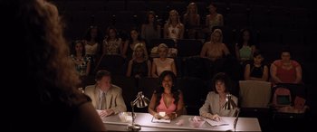 Movie still from “Dumplin'” (2018), directed by Anne Fletcher – A group of people sitting at a table in front of an audience; Wide shot, High angle
