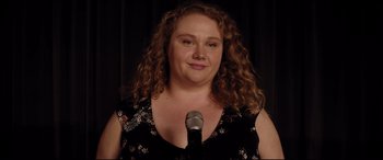 Movie still from “Dumplin'” (2018), directed by Anne Fletcher – A woman standing in front of a microphone with curly hair; Close Up shot, Over the shoulder angle
