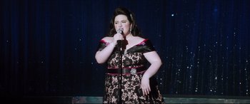 Movie still from “Dumplin'” (2018), directed by Anne Fletcher – A woman in a black and red dress singing into a microphone; Medium shot, High angle