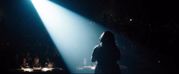 Movie still from “Dumplin'” (2018), directed by Anne Fletcher – A person standing on a stage in the middle of a dark room; Wide shot, High angle