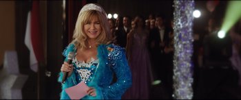 Movie still from “Dumplin'” (2018), directed by Anne Fletcher – A woman in a blue dress holding a microphone in front of a group of people; Medium shot, Over the shoulder angle