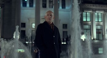 Movie still from “Elegy” (2008), directed by Isabel Coixet – A bald man in a black coat standing in front of a fountain; Medium shot, Low angle