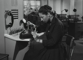 Movie still from “Elevator to the Gallows” (1958), directed by Louis Malle – A woman in a black dress is working on an object in an office setting; Medium shot, High angle