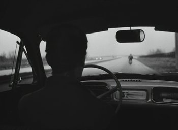Movie still from “Elevator to the Gallows” (1958), directed by Louis Malle – A man driving a car on a road with a motorcycle in the background; Wide shot, Over the shoulder angle