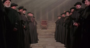 Movie still from “Elizabeth” (1998), directed by Shekhar Kapur – A group of men standing in front of a chair; Wide shot, High angle