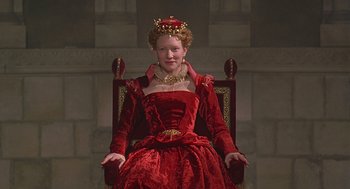 Movie still from “Elizabeth” (1998), directed by Shekhar Kapur – A woman in a red dress sitting in a chair; Medium shot, Low angle