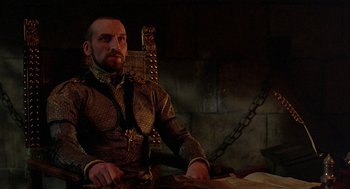 Movie still from “Elizabeth” (1998), directed by Shekhar Kapur – A man sitting at a table with a sword in his hand; Medium shot, Low angle