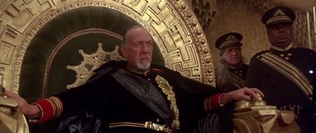 Movie still from “Dune” (1984), directed by David Lynch – An older man in a military uniform sitting in front of an ornate wall; Medium shot, Low angle