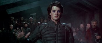 Movie still from “Dune” (1984), directed by David Lynch – A man in a black jacket standing in front of a group of people; Medium shot, Low angle