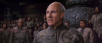 Movie still from “Dune” (1984), directed by David Lynch – A man with a bald head and blue eyes is standing in front of a group of people; Close Up shot, Over the shoulder angle