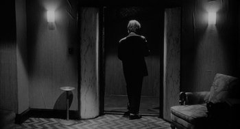Movie still from “Eraserhead” (1977), directed by David Lynch – A man in a suit standing in front of an elevator; Wide shot, Over the shoulder angle