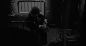 Movie still from “Eraserhead” (1977), directed by David Lynch – A person sitting on a bed looking at a phone; Medium shot, High angle