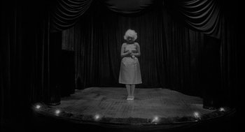 Movie still from “Eraserhead” (1977), directed by David Lynch – A woman standing on a stage in front of a curtain; Wide shot, Low angle