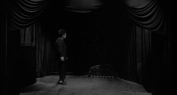 Movie still from “Eraserhead” (1977), directed by David Lynch – A man standing in front of an animal on a stage; Wide shot, High angle