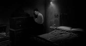 Movie still from “Eraserhead” (1977), directed by David Lynch – A man standing next to a bed in a room; Medium shot, High angle
