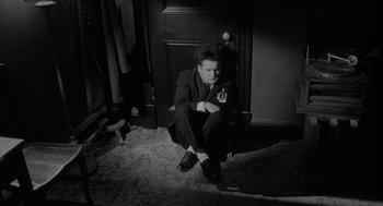 Movie still from “Eraserhead” (1977), directed by David Lynch – A black and white photo of a man smoking a cigarette in front of an open door; Medium shot, High angle