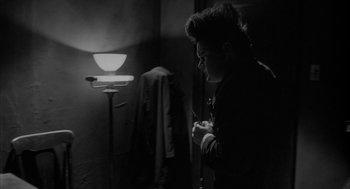 Movie still from “Eraserhead” (1977), directed by David Lynch – A man standing next to a lamp in a dark room; Medium shot, Over the shoulder angle