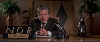 Movie still from “Escape from the Planet of the Apes” (1971), directed by Don Taylor – An older man sitting at a table in front of microphones; Medium shot, Low angle