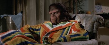 Movie still from “Escape from the Planet of the Apes” (1971), directed by Don Taylor – A gorilla is sitting in a chair wearing a colorful robe; Medium shot, High angle