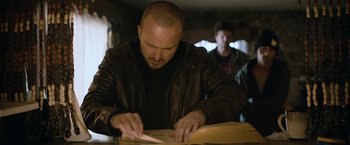 Movie still from “El Camino: A Breaking Bad Movie” (2019), directed by Vince Gilligan – A man wearing a leather jacket is cutting paper; Medium shot, Over the shoulder angle