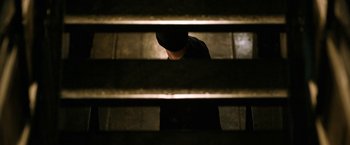 Movie still from “El Camino: A Breaking Bad Movie” (2019), directed by Vince Gilligan – A man standing in a dark room next to some stairs; Medium shot, Overhead angle