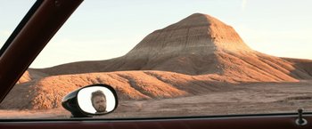 Movie still from “El Camino: A Breaking Bad Movie” (2019), directed by Vince Gilligan – A man's reflection in the side view mirror of a car; Wide shot, Over the shoulder angle