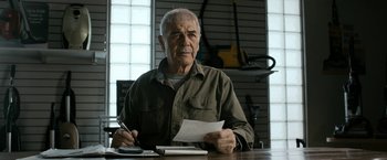 Movie still from “El Camino: A Breaking Bad Movie” (2019), directed by Vince Gilligan – An older man sitting at a table with a pen in his hand; Close Up shot, Low angle