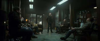Movie still from “El Camino: A Breaking Bad Movie” (2019), directed by Vince Gilligan – A group of people sitting in a room with one man standing up; Wide shot, Over the shoulder angle