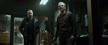 Movie still from “El Camino: A Breaking Bad Movie” (2019), directed by Vince Gilligan – Two men standing next to each other in a dark room; Medium shot, Low angle