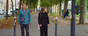 Movie still from “Everybody Loves Jeanne” (2022), directed by Céline Devaux – A woman standing on a sidewalk next to trees; Wide shot, Low angle