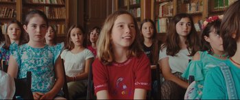 Movie still from “Everybody Loves Jeanne” (2022), directed by Céline Devaux – A group of young girls sitting in a room; Medium shot, Low angle