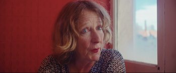 Movie still from “Everybody Loves Jeanne” (2022), directed by Céline Devaux – An older woman with blonde curly hair and blue eyes looking at the camera; Close Up shot, Over the shoulder angle