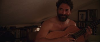 Movie still from “Everybody Loves Jeanne” (2022), directed by Céline Devaux – A man with a beard holding a guitar in a room; Medium shot, Over the shoulder angle
