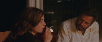 Movie still from “Everybody Loves Jeanne” (2022), directed by Céline Devaux – A woman smoking a cigarette while sitting at a table in front of a window; Medium shot, Over the shoulder angle
