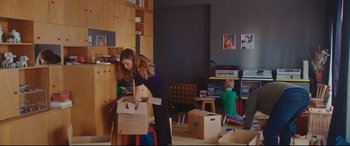 Movie still from “Everybody Loves Jeanne” (2022), directed by Céline Devaux – A woman holding a box in a living room; Wide shot, Over the shoulder angle