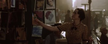 Movie still from “Evita” (1996), directed by Alan Parker – A woman in a brown and white dress is holding something up; Medium shot, Over the shoulder angle