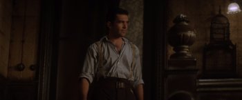 Movie still from “Evita” (1996), directed by Alan Parker – A man standing in a dark room wearing suspenders; Medium shot, Over the shoulder angle