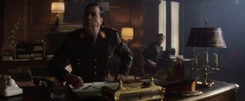 Movie still from “Evita” (1996), directed by Alan Parker – A man in a military uniform sitting at a table; Medium shot, Low angle