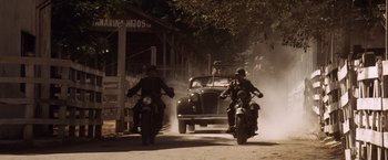 Movie still from “Evita” (1996), directed by Alan Parker – Two men on motorcycles driving down a dirt road past an old car; Wide shot, Low angle
