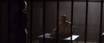 Movie still from “Evita” (1996), directed by Alan Parker – A man sitting at a table in front of a jail cell; Medium shot, Over the shoulder angle